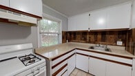 Located at 174 Diamond St in New Haven, Connecticut, this single-floor home offers a straightforward and functional layout with clearly defined living spaces. The kitchen is equipped with white cabinetry, light-toned countertops, and a distinctive wood-paneled backsplash that adds a rustic contrast to the otherwise neutral palette. A gas range, refrigerator, and double sink are positioned to maximize workspace efficiency, while a window above the counter allows natural light to filter in with views of surrounding greenery. The flooring in the kitchen consists of light-colored ceramic tile, providing a clean and durable surface. Adjacent living areas are finished with wall-to-wall carpet in a warm taupe tone, creating a comfortable and quiet atmosphere. The living room connects directly to the kitchen through a doorway, offering an open sense of flow between the two spaces. Bedrooms feature the same carpeted flooring and are painted in soft gray tones, with multiple windows that invite ample natural light and outdoor views. Crown molding and textured ceilings are consistent throughout the home, adding subtle architectural detail. The bathroom is finished with classic white square wall tiles, a tiled tub surround, a vanity with an undermount sink, and a window that provides ventilation and natural light.