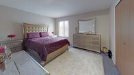 This single-floor residence offers a thoughtfully arranged layout with distinct living, dining, sleeping, and utility spaces. The living room serves as the central gathering area, featuring plush carpeting, a large sliding glass door with vertical blinds that allows generous natural light and wooded outdoor views, and wall-mounted television space. A tufted sofa and mirrored accent furnishings complement the room's warm, neutral tones.

Adjacent to the living room, the dining area accommodates a round glass-top table beneath an elegant crystal chandelier, with a window providing additional natural light. The galley-style kitchen connects directly to the dining space and features oak cabinetry, light stone-patterned countertops, white appliances, and wood-look vinyl flooring that extends into the dining area.

The primary bedroom is carpeted and offers ample space, with a wide sliding-door closet and a window with blinds. The second bedroom, similarly carpeted, features its own sliding-door closet and a window, and is well-suited as a children's room.

The full bathroom includes a tiled soaking tub and shower combination, an oak vanity with a light countertop, a large framed mirror, and wood-look vinyl flooring, offering a clean and functional design.