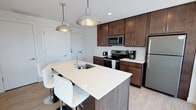 Located at 2202 S Kinnickinnic, this single-floor unit showcases a contemporary design with clean lines, light wood-tone flooring, and crisp white walls throughout. The kitchen is a standout feature, equipped with rich dark-stained cabinetry, white quartz countertops, stainless steel appliances, and a spacious island with an integrated sink and bar seating. Industrial-style pendant lights add character above the island, creating a functional yet stylish workspace.

The living area is bright and airy, benefiting from large floor-to-ceiling windows and a sliding glass door that opens to a private balcony overlooking a green outdoor space. A separate dining nook features oversized windows framing lively street views of the surrounding neighborhood.

The bedrooms are well-proportioned, with large windows that invite abundant natural light and offer tree-lined outdoor views. Soft carpet underfoot and roller shades provide comfort and privacy. The bathroom maintains the unit's modern aesthetic with light tile flooring, a dark-stained vanity, white countertop, and a walk-in shower area. Throughout the space, the consistent use of neutral tones, modern hardware, and thoughtful layout create a cohesive, polished living environment.
