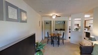 This single-floor space presents an open and functional layout that seamlessly integrates the living and dining areas. The living area is anchored by a large flat-screen television and accented with a framed wall piece, while a potted plant adds a touch of greenery. Neutral-toned carpet flows throughout, contributing to a warm and cohesive feel underfoot.

A ceiling fan with a built-in light fixture is centrally positioned overhead, providing both air circulation and ambient lighting. The textured ceiling adds subtle visual interest to the space.

The dining area features a round wooden table surrounded by four chairs, creating an intimate setting for meals. Behind the dining area, a set of open shelving units with bifold-style doors provides practical storage and display space, housing books and small decorative items.

A pass-through opening connects the main living and dining area to the adjacent kitchen, which is partially visible and appears bright and functional. This architectural detail enhances the open feel of the overall space, allowing light to travel freely between rooms.