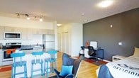 This modern single-floor unit showcases a thoughtfully designed open-concept layout that seamlessly integrates the kitchen, dining, and living areas into one cohesive space. The kitchen is outfitted with stainless steel appliances, including a full-sized refrigerator, and features light wood cabinetry, a sleek countertop, and a built-in sink. A breakfast bar with stylish blue bar stools serves as a natural dividing point between the kitchen and the main living area, making it ideal for casual dining or entertaining. The living space is warmed by light hardwood flooring that runs throughout, creating a bright and inviting atmosphere. A bold dark accent wall anchors the workspace corner, which is furnished with a desk, an ergonomic office chair, and storage, offering a dedicated area for productivity. Comfortable lounge chairs with accent pillows complete the seating arrangement, adding a relaxed, contemporary feel. Track lighting in the kitchen and flush-mount ceiling fixtures provide ample illumination throughout. A set of closet doors hints at additional storage solutions, enhancing the functional appeal of this well-appointed space.