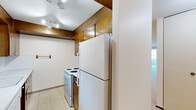 This two-bedroom, one-bathroom residence in Tukwila, Washington offers a practical and well-organized single-floor layout. The kitchen features warm wood cabinetry extending to the upper walls, white tile flooring, a coil-burner electric range, a full-size refrigerator, and a white countertop with an integrated sink — providing solid functionality for everyday cooking. The two bedrooms are carpeted and bright, each benefiting from natural light filtering through vertical-blind windows. Both bedrooms include sliding-door closets with gold-toned hardware, offering generous storage. Baseboard heating units run beneath the windows in the bedrooms, ensuring comfortable warmth. The full bathroom is cleanly appointed with a tub/shower combination, a wall-mounted sink, and tile flooring. Hallways connect the rooms efficiently, and a balcony extends the living space outdoors. The overall color palette is neutral throughout — cream walls, light carpet, and wood-toned doors and trim — creating a clean, move-in-ready feel. The layout, as seen in the dollhouse view, makes efficient use of the available floor area, with logical flow between the living, sleeping, and kitchen spaces.