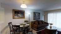 This three-floor space offers a thoughtfully curated interior defined by classic elegance and warm, inviting character. The living and dining area exemplifies the overall aesthetic, where a richly upholstered sofa with ornate fringe detailing anchors the sitting area alongside a bold patterned area rug. A round dark-wood dining table with intricate wrought-iron chairs creates an intimate dining setting, complete with place settings and a decorative centerpiece. Ambient lighting is provided through a combination of a central dome pendant light and tall floor lamps that cast a soft, warm glow throughout the space. The walls are adorned with large, framed oil-style paintings — including a detailed still life with fruit and a scenic landscape — adding a gallery-like sophistication to the rooms. The textured ceiling and neutral wall tones serve as a subtle backdrop that allows the furnishings and artwork to take center stage. Spanning three floors, the space provides a generous sense of vertical scale and room for varied living functions across each level. The overall ambiance is warm, traditionally styled, and richly detailed, making it a space that feels both lived-in and carefully appointed.