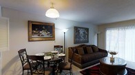This three-floor space offers a thoughtfully curated interior defined by classic elegance and warm, inviting character. The living and dining area exemplifies the overall aesthetic, where a richly upholstered sofa with ornate fringe detailing anchors the sitting area alongside a bold patterned area rug. A round dark-wood dining table with intricate wrought-iron chairs creates an intimate dining setting, complete with place settings and a decorative centerpiece. Ambient lighting is provided through a combination of a central dome pendant light and tall floor lamps that cast a soft, warm glow throughout the space. The walls are adorned with large, framed oil-style paintings — including a detailed still life with fruit and a scenic landscape — adding a gallery-like sophistication to the rooms. The textured ceiling and neutral wall tones serve as a subtle backdrop that allows the furnishings and artwork to take center stage. Spanning three floors, the space provides a generous sense of vertical scale and room for varied living functions across each level. The overall ambiance is warm, traditionally styled, and richly detailed, making it a space that feels both lived-in and carefully appointed.