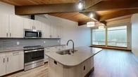 Located at 1940 W Riverstone Drive in Coeur d'Alene, Idaho, this single-floor residence combines contemporary finishes with warm architectural character. The open-concept kitchen is a standout feature, showcasing exposed wood beam ceilings with tongue-and-groove wood paneling, a large quartz-topped island with an undermount sink, light wood cabinetry, a mosaic tile backsplash, and a full suite of stainless steel appliances. Geometric pendant lights add a modern touch overhead. The living and dining areas extend naturally from the kitchen, benefiting from large windows that invite abundant natural light and offer views of the surrounding Coeur d'Alene landscape. Light hardwood-style flooring flows throughout the main living spaces, complemented by rich dark baseboards and door trim. The bedroom is generously sized, featuring plush carpet, exposed beam accents, a sliding glass door to an outdoor balcony, and a spacious closet with sliding panel doors. A secondary room with wide windows and hardwood flooring provides flexible use as an office or additional bedroom. The bathroom is finished with large-format tile flooring and a sleek walk-in shower enclosed by black-framed glass doors, contributing a clean, modern aesthetic throughout.