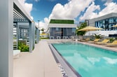 Located at 51 NW 28th Street in Miami's vibrant Wynwood district, this rooftop amenity deck offers a resort-inspired outdoor experience elevated above the city. The centerpiece is a generously sized swimming pool with crystal-clear turquoise water, finished with clean tile edging and surrounded by a spacious travertine-style pool deck. Rows of modern yellow-cushioned sun loungers and striped market umbrellas line the pool's perimeter, providing ample space for relaxation and sunbathing under the South Florida sky.

Architecturally, the deck incorporates sleek pergola structures with louvered panels that offer shaded seating areas and a sense of privacy. Lush tropical landscaping—including tall palm trees, banana plants, and a variety of tropical foliage—frames the space, creating a garden-like atmosphere that softens the urban setting. A dedicated lawn area with manicured artificial turf and additional lounge seating extends along one side of the deck, offering a casual, open-air retreat.

A glass-fronted clubroom or amenity pavilion anchors one end of the rooftop, providing an interior space that seamlessly connects to the outdoor deck. The space commands open panoramic views of the Wynwood and greater Miami skyline, making it an ideal setting for both relaxation and social gatherings throughout the day.