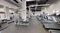 Located within the iconic Terminal Tower in Cleveland, Ohio, this fitness center delivers a comprehensive workout facility across a single, expansive floor. The space features an industrial-modern aesthetic, with exposed ductwork and black-painted ceiling structures contrasting against crisp white walls and bright linear LED lighting, creating a clean and energizing atmosphere.

The cardio zone is well-appointed with a long row of treadmills, elliptical trainers, stationary bikes, and rowing machines, all equipped with individual display screens. Multiple wall-mounted flat-screen televisions are positioned throughout the facility for easy viewing from virtually any workout station.

A dedicated strength training area houses a variety of cable and selectorized machines, along with a free weight section featuring kettlebells, dumbbells, and adjustable benches. Additional functional training equipment, including medicine balls, rounds out the offerings.

Natural light filters in through several windows positioned along the walls, brightening the space and providing views of the surrounding urban environment. Durable rubber flooring spans the entire facility, providing safety and comfort underfoot. The open floor plan and strategic equipment layout ensure ample circulation space, making the Terminal Tower Fitness Center a well-organized and fully equipped destination for all fitness levels.
