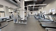 Located within the iconic Terminal Tower in Cleveland, Ohio, this fitness center delivers a comprehensive workout facility across a single, expansive floor. The space features an industrial-modern aesthetic, with exposed ductwork and black-painted ceiling structures contrasting against crisp white walls and bright linear LED lighting, creating a clean and energizing atmosphere.

The cardio zone is well-appointed with a long row of treadmills, elliptical trainers, stationary bikes, and rowing machines, all equipped with individual display screens. Multiple wall-mounted flat-screen televisions are positioned throughout the facility for easy viewing from virtually any workout station.

A dedicated strength training area houses a variety of cable and selectorized machines, along with a free weight section featuring kettlebells, dumbbells, and adjustable benches. Additional functional training equipment, including medicine balls, rounds out the offerings.

Natural light filters in through several windows positioned along the walls, brightening the space and providing views of the surrounding urban environment. Durable rubber flooring spans the entire facility, providing safety and comfort underfoot. The open floor plan and strategic equipment layout ensure ample circulation space, making the Terminal Tower Fitness Center a well-organized and fully equipped destination for all fitness levels.