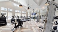 Located at 5948 Aretha Place in Virginia Beach, VA, this modern fitness center spans a generously sized single-floor layout divided into two functional workout zones. The space is defined by its dramatic vaulted ceilings in the main area, which are accentuated by striking black pendant lights that add an industrial-chic contrast to the bright, white interior. Large windows line the walls throughout, flooding both areas with natural light and creating an open, energizing atmosphere.

The primary workout area features an impressive array of cardio equipment, including multiple treadmills, ellipticals, air bikes, and stationary cycles, all arranged along a wall of floor-to-ceiling mirrors that enhance the sense of space. A wall-mounted flat-screen TV provides entertainment during workouts. The flooring throughout features a distinctive herringbone-patterned tile in neutral tones of beige, tan, and gray, lending a polished, contemporary aesthetic.

The secondary area, accessible through sleek black-framed glass doors, is dedicated to strength training. It includes a multi-station cable machine, free weights, kettlebells, medicine balls, and weight benches. Additional mirrored walls and mounted TVs complete this zone. Together, the two areas provide a comprehensive, well-appointed fitness facility suited for a full range of exercise routines.