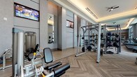 This fitness center offers a spacious, thoughtfully designed environment suited for a wide range of workout routines. The space features striking herringbone-patterned flooring in warm neutral tones, which complements the soft grey walls and contributes to the overall polished aesthetic. Elevated ceilings with recessed cove lighting add an airy, upscale feel to the room, while large windows along one wall allow abundant natural light to fill the space.

The cardio zone is well-equipped with multiple treadmills, elliptical machines, and stationary bikes, all positioned to take advantage of the natural light and window views. A ballet barre runs along a mirrored wall in this section, supporting stretching and functional movement exercises. Full-length mirrors are strategically placed throughout to enhance spatial openness and support form-checking during workouts.

The strength training area features a robust selection of equipment, including cable machines, multi-functional rig systems with TRX suspension trainers, adjustable benches, and a comprehensive dumbbell rack. Medicine balls, kettlebells, and weight plates are neatly organized for easy access. Wall-mounted flat-screen televisions are positioned throughout the space, ensuring entertainment is visible from multiple workout stations. Ceiling fans provide additional airflow, rounding out this well-equipped and elegantly finished fitness facility.