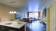 This single-floor unit showcases an open-concept design that integrates the kitchen, dining, and living areas into one cohesive space. The kitchen features a granite-topped island with pendant lighting overhead, providing both a functional workspace and a natural dividing point between cooking and entertaining areas. Dark hardwood-style flooring runs throughout, adding warmth and continuity to the layout.

The dining area sits centrally within the space, furnished with a dark wood table and upholstered chairs, positioned beneath contemporary pendant lights. A large piece of colorful wall art adds a vibrant focal point to this zone. An accent wall in a warm terracotta hue frames the living area, drawing the eye toward the expansive picture window at the far end of the room. This large window floods the space with natural light and offers views of the surrounding greenery outdoors.

The living area is generously proportioned, comfortably accommodating a full sofa arrangement alongside a dedicated workspace. The overall ambiance is modern and welcoming, with a balanced combination of natural light, thoughtful accent colors, and cohesive finishes throughout.