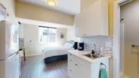 This single-level studio unit offers an efficient, open-concept layout that thoughtfully combines living and functional spaces within a compact footprint. The main area features warm-toned walls, durable wood-look flooring, and elevated ceilings that enhance the sense of airiness throughout the space.

The sleeping area is positioned to take advantage of natural light from large windows, which frame views of the surrounding neighborhood and allow sunlight to fill the room throughout the day. A wall-mounted television and a small media stand provide a comfortable entertainment setup opposite the bed.

The kitchenette is seamlessly integrated into the main living space and is outfitted with white cabinetry — both upper and lower — topped with light countertops and a stainless steel sink. A mosaic tile backsplash adds a decorative detail to this functional area, which also accommodates a coffee maker and basic prep essentials.

A dedicated bathroom is accessible from the main space and includes a toilet with overhead shelving for storage, as well as a towel bar. The bathroom's neutral palette maintains the clean, cohesive aesthetic found throughout the unit.

Overall, the space presents a tidy, modern ambiance well-suited for single occupancy, combining practical amenities with a straightforward, functional design.