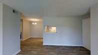 This single-floor space offers a clean, open layout designed for comfortable everyday living. The living and dining areas flow seamlessly together, unified by durable wood-look plank flooring that runs throughout. Neutral light gray walls create a bright, airy atmosphere that complements the understated aesthetic of the space.

The dining area is anchored by a modest chandelier, providing warm ambient lighting and a subtle decorative focal point. A standout architectural feature is the kitchen pass-through window, which connects the kitchen to the main living area. This opening is framed by a stone or granite-style ledge and provides a glimpse into the kitchen, where gray cabinetry, a classic subway tile backsplash, and under-cabinet lighting are visible. The pass-through enhances the sense of openness while maintaining a degree of separation between the spaces.

White baseboards throughout offer a crisp contrast to the flooring and walls, adding a polished finishing detail. The overall ambiance is clean, modern, and move-in ready, with a neutral palette that provides a versatile backdrop for a variety of interior styles.