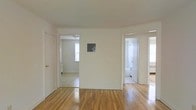 This single-floor space presents a straightforward and functional layout characterized by clean white walls and warm hardwood flooring that runs throughout. The main living area serves as a central hub, with doorways opening to several adjoining rooms. To the left, a bedroom is visible, benefiting from natural light filtering through a window and featuring its own flooring. To the right, a bathroom is accessible, appearing tiled and equipped with standard fixtures including what appears to be a bathtub or shower. A small electrical panel is mounted on the central wall, a practical detail integrated discreetly into the space. The hardwood floors throughout add warmth and continuity, reflecting light and contributing to an overall bright atmosphere. The white walls and neutral palette keep the space feeling open and adaptable. The layout is efficient, with each room clearly defined yet connected, making good use of the available footprint. Overall, the space offers a clean, uncluttered environment well-suited for straightforward, comfortable living.