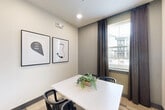 Located at 698 Basilica Bay Drive in Spring, TX, this conference room offers a focused and functional environment for small meetings and collaborative sessions. The room is designed with a clean, modern aesthetic, featuring neutral gray and white walls that create a calm, professional atmosphere. A central white rectangular table is surrounded by black accent chairs, providing seating for a small group. A wall-mounted flat-screen TV serves as a key feature, ideal for presentations or video conferencing. Natural light filters into the space through a window fitted with horizontal blinds and dressed with rich dark curtains, adding warmth to the otherwise crisp interior. The floor is covered in patterned carpet tiles in a neutral, multi-toned design that complements the contemporary style. Two framed black-and-white art pieces adorn the walls, lending a thoughtful, curated touch to the space. The room is accessed through a gray-framed glass-paneled door, maintaining visual connectivity with the adjacent area while offering privacy. A subtle greenery centerpiece on the table adds a refreshing organic element. Recessed lighting and a wall-mounted security camera complete the practical amenities of this well-appointed conference room.