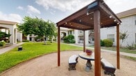 This expansive outdoor courtyard serves as a central communal green space, enclosed by two-story brick buildings featuring distinctive arched entryways, stucco accents, and warm earth-toned trim. The architecture presents a consistent, cohesive aesthetic with covered walkways, wrought iron balcony railings, and stairwells connecting the ground and upper levels.

The courtyard itself is characterized by well-kept, lush green lawns that stretch across the length of the space, punctuated by mature shade trees with broad canopies that provide natural cooling and visual interest. Carefully maintained landscaping beds line the building perimeters, featuring a variety of ornamental shrubs, flowering bushes, and decorative ground cover that add color and texture throughout the seasons.

A central pergola with an attached picnic table offers a shaded gathering spot at the heart of the courtyard, complemented by additional stone benches positioned along the pathways for informal seating. Concrete walkways crisscross the grounds, providing convenient pedestrian access to all building entrances. A dedicated playground area at one end of the courtyard includes play structures set within sand-filled zones, offering recreational space for younger residents. Outdoor lighting posts are distributed throughout, ensuring the courtyard remains functional and welcoming into the evening hours.