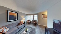 This single-floor space features a well-appointed living area that combines modern design with a comfortable, functional layout. The room is finished with wood-style plank flooring throughout, adding warmth and a clean aesthetic to the space. A textured popcorn ceiling adds dimension overhead, while neutral gray walls provide a versatile backdrop that complements a variety of décor styles.

Large windows fitted with horizontal blinds allow generous amounts of natural light to fill the room, creating a bright and welcoming atmosphere. The living area is spacious enough to accommodate a full sofa arrangement alongside a dedicated seating nook near the windows, making it ideal for both relaxing and entertaining. A glass-top coffee table over a plush area rug anchors the main seating area, enhancing the open feel of the space.

Framed artwork on the walls adds visual interest, while recessed lighting and a freestanding lamp provide flexible illumination options. The overall ambiance is contemporary and inviting, offering a balanced blend of style and practicality in a single cohesive living environment.