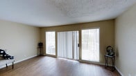This two-floor space offers a clean, functional layout with notable design details throughout. The main living area showcases warm hardwood-style flooring that runs seamlessly across the room, complementing the neutral beige walls that create a welcoming, understated atmosphere. A textured ceiling adds subtle character overhead.

A standout feature of the living area is the generous wall of windows and sliding glass doors, which span nearly the full width of the exterior wall. These doors provide direct access to an outdoor deck or balcony, flooding the interior with natural light and offering views of the surrounding outdoor environment. Vertical blinds on the sliding doors allow for flexible light and privacy control throughout the day.

The overall ambiance of the space is open and airy, with the abundance of natural light enhancing the sense of spaciousness. The simple, neutral color palette throughout provides a versatile backdrop adaptable to a variety of furnishing styles. Spanning two floors, the property offers additional living space beyond what is visible, providing ample room for a range of living arrangements.