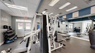 Located at 3343 Glade Creek Blvd NE in Roanoke, Virginia, this fitness center offers a fully equipped exercise room designed to support a wide range of workout routines. The space features a drop ceiling with recessed fluorescent lighting, rubber-mat flooring for safety and durability, and a calming blue-and-gray color palette that creates a clean, modern aesthetic. Motivational wall art and the phrase "Your Only Limit Is You" add an encouraging atmosphere throughout.

The cardio section is well-stocked with treadmills, elliptical machines, a stair climber, and a stationary bike, all positioned near windows that allow natural light and outdoor views to enhance the workout experience. A wall-mounted flat-screen TV provides entertainment for cardio sessions. The strength training area includes a multi-station cable machine, weight benches, barbells, and a full rack of dumbbells, catering to both beginners and experienced athletes. Large wall mirrors in the free weights section allow users to monitor their form effectively.

A separate room accessible through glass French doors appears to serve as a restroom or utility space. The fitness center also benefits from direct exterior access, making it convenient for residents. Overall, the space is thoughtfully organized to maximize functionality while maintaining a welcoming and well-maintained environment.