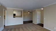 This single-floor residence offers a thoughtfully designed open-concept layout that promotes a sense of spaciousness and easy flow between living areas. The main living space is finished with plush carpeting and transitions seamlessly to dark hardwood-style flooring, creating a clear visual distinction between zones. Crown molding runs throughout, lending an elegant, polished character to the interior. A ceiling fan with a light fixture is centrally positioned in the living area, providing both comfort and ambient lighting. The kitchen is partially separated by a half-wall pass-through, which maintains an open connection to the living space while providing a functional counter surface. Inside the kitchen, white cabinetry offers generous storage, and a built-in microwave is visible alongside the upper cabinets. The neutral, warm-toned walls throughout complement the crisp white trim and baseboards, creating a cohesive and inviting atmosphere. A paneled interior door provides access to an additional room, adding to the versatility of the floor plan. The overall design balances practicality with classic detailing, making the space feel both functional and comfortable.