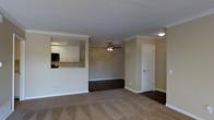 This single-floor residence offers a thoughtfully designed open-concept layout that promotes a sense of spaciousness and easy flow between living areas. The main living space is finished with plush carpeting and transitions seamlessly to dark hardwood-style flooring, creating a clear visual distinction between zones. Crown molding runs throughout, lending an elegant, polished character to the interior. A ceiling fan with a light fixture is centrally positioned in the living area, providing both comfort and ambient lighting. The kitchen is partially separated by a half-wall pass-through, which maintains an open connection to the living space while providing a functional counter surface. Inside the kitchen, white cabinetry offers generous storage, and a built-in microwave is visible alongside the upper cabinets. The neutral, warm-toned walls throughout complement the crisp white trim and baseboards, creating a cohesive and inviting atmosphere. A paneled interior door provides access to an additional room, adding to the versatility of the floor plan. The overall design balances practicality with classic detailing, making the space feel both functional and comfortable.