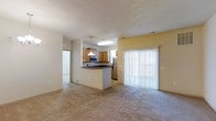 This single-floor residence offers an open-concept layout that seamlessly connects the living and kitchen areas, creating a spacious and functional environment. The kitchen is well-appointed with light wood cabinetry, dark countertops, stainless steel appliances including a microwave and refrigerator, and is illuminated by a stylish pendant light above. A peninsula-style breakfast bar serves as a natural divider between the kitchen and the carpeted living area, providing additional prep space and casual seating options. The living room is bathed in natural light streaming through a large sliding glass door dressed with vertical blinds, offering easy access to the outdoors. Neutral-toned carpeting extends throughout the living space, contributing to a warm and inviting atmosphere. The soft, textured ceiling and warm yellow-beige walls add to the cozy character of the interior. A hallway visible from the main living area suggests additional rooms beyond, providing practical separation of living zones within the single-story floor plan. Overall, the space is designed for comfortable everyday living with a clean, functional aesthetic.