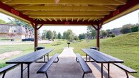 Located at 111 Whitsett Rd in Nashville, Tennessee, this outdoor pavilion offers a well-structured, open-air gathering space designed for community use. The pavilion features a bold red and yellow wooden roof structure with exposed beam rafters, supported by sturdy red posts that frame sweeping views of the surrounding landscape on all sides. A ceiling fan is mounted within the roofline, providing airflow during warm Tennessee days.

The concrete floor accommodates multiple dark, heavy-duty picnic tables with attached bench seating, providing ample space for groups to gather, dine, or socialize. Trash receptacles are conveniently positioned throughout the pavilion for easy access.

The pavilion opens onto expansive, well-maintained grassy grounds dotted with mature trees, including weeping willows, that contribute to a lush, park-like atmosphere. A paved walkway extends from the pavilion into the grounds, where additional outdoor amenities such as grills and benches are visible. Views from within the pavilion extend in multiple directions, capturing open lawn areas, wooded backdrops, and neighboring structures. The open-sided design ensures abundant natural light and ventilation throughout the space, making it a functional and inviting venue for outdoor gatherings in any direction.