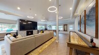 Located at 15195 SW Walker Rd in Beaverton, Oregon, this single-story community clubhouse offers a thoughtfully designed gathering space ideal for socializing and recreation. The open-concept layout is anchored by a spacious lounge area featuring a large sectional sofa, a marble coffee table, and a striking herringbone-tiled fireplace with a wall-mounted television above. Dramatic circular pendant lights hang from the soaring ceilings, adding a contemporary flair throughout the main area. Adjacent to the lounge, a dedicated game room houses a pool table and a full-length shuffleboard table, providing ample entertainment options. A cozy seating nook with swivel chairs, a decorative area rug, and curated wall art creates an inviting conversation area, bathed in natural light from large windows overlooking the outdoor pool and greenery. A compact bar-height workspace with stools sits beside panoramic windows, offering views of the surrounding community and pool area. The fully equipped kitchenette features stainless steel appliances, a mosaic tile backsplash, light stone countertops, and a dining table for casual meals. Light wood-look flooring flows throughout, tying together the bright, neutral palette and modern aesthetic of this versatile community space.