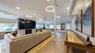 Located at 15195 SW Walker Rd in Beaverton, Oregon, this single-story community clubhouse offers a thoughtfully designed gathering space ideal for socializing and recreation. The open-concept layout is anchored by a spacious lounge area featuring a large sectional sofa, a marble coffee table, and a striking herringbone-tiled fireplace with a wall-mounted television above. Dramatic circular pendant lights hang from the soaring ceilings, adding a contemporary flair throughout the main area. Adjacent to the lounge, a dedicated game room houses a pool table and a full-length shuffleboard table, providing ample entertainment options. A cozy seating nook with swivel chairs, a decorative area rug, and curated wall art creates an inviting conversation area, bathed in natural light from large windows overlooking the outdoor pool and greenery. A compact bar-height workspace with stools sits beside panoramic windows, offering views of the surrounding community and pool area. The fully equipped kitchenette features stainless steel appliances, a mosaic tile backsplash, light stone countertops, and a dining table for casual meals. Light wood-look flooring flows throughout, tying together the bright, neutral palette and modern aesthetic of this versatile community space.