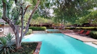 This expansive outdoor pool area offers a resort-like atmosphere defined by its thoughtfully designed landscaping and well-appointed amenities. The generously sized swimming pool features clear turquoise water and accommodates both shallow and deep ends, with depth markers clearly visible along the pool's edge. Stone-clad retaining walls and raised planting beds add architectural character, seamlessly integrating the natural surroundings with the hardscaped pool deck. A decorative water fountain serves as a focal point near one end of the pool, providing a soothing ambiance. Mature oak, willow, and crepe myrtle trees create a lush, shaded canopy overhead, offering natural relief from the sun while enhancing the sense of privacy and tranquility. A wooden pergola provides additional shaded seating and lounging space, complemented by red lounge chairs and patio furniture with umbrellas positioned around the deck. The surrounding brick and stone pool deck offers generous space for relaxation. Adjacent to the pool, a dark wood-clad community building with a striking pitched roofline provides covered outdoor gathering areas and picnic seating. The overall ambiance is peaceful and resort-inspired, making this pool area an exceptional outdoor retreat.
