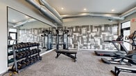 Located at 5532 38th St S in Fargo, ND, this fitness center is a thoughtfully designed, single-floor workout facility offering a comprehensive range of exercise equipment within a compact yet efficiently utilized space. The room features neutral-toned carpet flooring throughout and is illuminated by recessed ceiling lights, contributing to a clean and focused workout environment. Exposed industrial-style ductwork runs along the ceiling, adding a modern, urban aesthetic to the space. A standout feature is the bold, large-scale black-and-white graphic accent wall that spans a significant portion of the room, providing a visually dynamic backdrop. Full-length mirrors along one wall enhance the sense of openness and allow users to monitor their form during exercise. The cardio area lines the windowed wall, where multiple treadmills, ellipticals, and a rowing machine are positioned in front of large windows that invite natural light and outdoor views. Wall-mounted flat-screen TVs are positioned for easy viewing from the cardio equipment. The strength training area is well-appointed with a multi-station cable machine, an adjustable weight bench, and a full rack of dumbbells. A water fountain and storage bench are also present, completing this practical and modern fitness facility.
