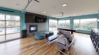 This dynamic single-floor amenity space is designed for social gatherings and relaxation, unified by bold teal accent walls and rich hardwood flooring throughout. The open-plan layout flows seamlessly from a fully equipped kitchen area to a spacious lounge, creating an inviting and functional environment.

The kitchen features stainless steel appliances, including a full-size refrigerator and microwave, dark wood cabinetry, a mosaic tile backsplash, and a generous white countertop bar with seating for multiple guests. Pendant lighting adds a polished touch overhead, while a wall-mounted television keeps the space entertained.

The main lounge area is anchored by a sleek, tile-surround fireplace topped with a large flat-screen TV, creating a natural focal point. Contemporary curved ottomans and modular seating arrangements provide comfortable gathering spots, while a billiards table offers additional entertainment. Industrial-style ceiling-mounted hardware and recessed lighting contribute to the space's modern, loft-inspired aesthetic.

Floor-to-ceiling glass panels with overhead garage-style doors open directly onto a furnished outdoor terrace, flooding the interior with natural light and offering sweeping city views. The terrace extends the living space outdoors with additional seating, making it ideal for social events or quiet relaxation while taking in the urban skyline.