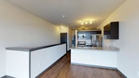 This single-floor space showcases a contemporary open-concept design that seamlessly integrates the living and kitchen areas. The kitchen is a standout feature, equipped with stainless steel appliances including a refrigerator, microwave, and range. Dark espresso cabinetry provides ample storage and contrasts sharply with the light granite-style countertops of the extended kitchen island, which also functions as a breakfast bar or prep space. Track lighting above the kitchen adds focused illumination, enhancing the modern aesthetic. The flooring throughout is a warm, dark hardwood-style material that anchors the space with visual depth and durability. A half-wall partition helps define the living area from a hallway corridor, adding architectural structure without fully closing off the open layout. The neutral wall tones and consistent ceiling height contribute to a clean, understated atmosphere. A dark-framed entry door is visible in the background, offering a cohesive design element. Overall, the space presents a functional and stylish environment suited for modern living, with thoughtful material choices and a well-organized layout.