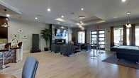 This contemporary communal lounge space is thoughtfully designed to balance comfort and sophistication. The centerpiece of the room is a sleek, floor-to-ceiling stone-clad accent wall that frames both a linear gas fireplace and a large wall-mounted television, creating a striking focal point that anchors the main seating area. Dark charcoal upholstered sofas and armchairs are arranged around a patterned area rug, providing an inviting space for relaxation and conversation. Light hardwood flooring extends throughout, lending warmth and continuity to the modern aesthetic. A tray ceiling detail overhead adds architectural depth, while recessed lighting and a stunning cluster pendant chandelier provide layered illumination. Adjacent to the main lounge area, a high-top bar seating section with sleek stools offers an alternate gathering space, ideal for casual socializing. Tall, dark-framed glass doors invite natural light and provide a visual connection to the outdoors. Lush tropical potted plants and a sculptural water feature introduce organic elements that soften the otherwise clean-lined, urban design. The overall ambiance is polished, modern, and welcoming.