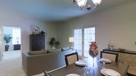 This single-floor space showcases an open-concept layout that seamlessly integrates a dining area and living room into one cohesive environment. The dining area is anchored by a dark wood table surrounded by matching upholstered chairs, set beneath an elegant chandelier with ornate metalwork that serves as a focal point overhead. A decorative glass vase filled with fruits and natural accents adds a warm, inviting centerpiece to the table.

The adjacent living area features a curved sectional sofa in neutral tones, complemented by a dark wood cabinet that provides both storage and display space for decorative accents. A floor lamp provides ambient lighting, contributing to the room's warm atmosphere. The walls are finished in a soft sage green, lending the space a calm and welcoming feel.

French doors with grid-pane detailing allow abundant natural light to flow into the space while offering views of the outdoors. The light-toned flooring ties the two areas together, enhancing the sense of openness throughout. Overall, the space balances functional living with tasteful décor.