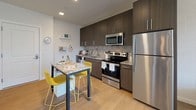 Located at 200 Pleasant St in Malden, MA, this single-floor residence offers a thoughtfully designed layout that balances modern aesthetics with functional living. The kitchen is a standout feature, equipped with dark espresso cabinetry extending to the ceiling, granite countertops, a tiled backsplash, and a full suite of stainless steel appliances including a refrigerator, range, microwave, and dishwasher. A granite-topped island provides additional prep and dining space, complemented by light hardwood-style flooring throughout. The living area is bathed in natural light through a large floor-to-ceiling window offering views of the surrounding urban streetscape, creating an open and airy atmosphere. The bedroom continues the bright, contemporary feel with oversized windows, neutral carpet, and clean white walls. The full bathroom features large-format tile on the floors and shower surround, a soaking tub, and a streamlined vanity with ample counter space. An adjacent walk-in closet adds practical storage. Throughout the unit, a neutral color palette and modern finishes create a cohesive and inviting ambiance suited to contemporary urban living.