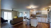 This single-floor residence showcases a thoughtfully designed open-concept layout that unifies the living and kitchen areas into one cohesive, functional space. Rich hardwood flooring flows throughout, lending warmth and continuity to the interior. The kitchen is well-appointed with crisp white cabinetry, granite-toned countertops, and a central island featuring bar-height seating — perfect for casual dining or entertaining. Pendant lighting above the island adds a stylish focal point, while under-cabinet and recessed lighting ensure the space is well-lit. The adjacent living area is generously proportioned, centered around a comfortable sectional sofa and accented with a ceiling fan that aids in year-round comfort. Natural light filters in through windows, brightening the neutral color palette of warm beiges and soft grays that unifies the space. A framed artwork piece anchors the living area's wall, adding a decorative touch. A doorway visible from the main living space leads into what appears to be a bedroom, hinting at a private retreat beyond the open common areas. Overall, the space offers a welcoming, practical, and attractively finished environment suited to comfortable everyday living.