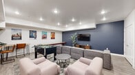 This dedicated entertainment and game room is thoughtfully designed for social gatherings and recreational use. The open, single-floor layout makes efficient use of its generous footprint, offering distinct zones for gaming, lounging, and casual dining. Recessed lighting throughout provides bright, even illumination across the entire space.

The gaming area is a central highlight, featuring a full-size foosball table and a long shuffleboard table positioned along one wall, both framed by a curated collection of classic movie posters — including Star Wars, Rocky, The Goonies, E.T., and The Princess Bride — that lend the room a fun, pop-culture character.

The lounge zone is anchored by a large sectional sofa in grey and complemented by blush-pink accent chairs arranged around a round coffee table, all set atop a geometric area rug over light wood-style flooring. A wall-mounted flat-screen TV sits against a bold navy blue accent wall, accompanied by a wooden media console and a tall indoor plant that adds a touch of greenery.

A bar-height dining area with hairpin-leg tables and industrial-style stools provides additional seating. Double panel doors offer easy access to and from the space. The overall ambiance is modern, vibrant, and inviting — ideal for entertaining groups of all sizes.