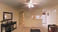 This single-floor residence features an open-concept layout that connects the living area to the kitchen, creating a cohesive and functional living space. The living area is finished with wall-to-wall carpeting in a neutral tone, contributing to a warm and comfortable ambiance. A ceiling fan with an integrated light fixture is centrally mounted, providing both air circulation and overhead illumination throughout the space.

The kitchen is partially open to the living area via a raised breakfast bar counter, offering a casual dining or serving surface while maintaining a visual connection between the two spaces. The kitchen is equipped with white appliances, including a refrigerator and microwave, and features wooden cabinetry that adds a classic touch. A louvered utility access panel is visible on the shared wall between the living and kitchen areas.

A hallway visible from the main living space leads to additional rooms, suggesting a practical layout designed for easy navigation. The textured ceiling and neutral wall tones throughout create a cohesive, understated aesthetic. The overall design prioritizes functionality while maintaining a welcoming, comfortable atmosphere suitable for everyday living.