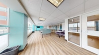This contemporary open-plan workspace is designed to support a variety of working and studying styles within a well-organized, single-floor layout. The space features light-toned hardwood-style flooring that runs throughout, contributing to a clean and welcoming atmosphere. A drop tile ceiling with recessed and panel lighting ensures consistent, even illumination across the entire floor.

Individual workstations equipped with teal acoustic privacy dividers are arranged throughout the main area, providing focused work zones without fully enclosing the space. A lounge area with plush purple armchairs and a small table offers a comfortable setting for informal meetings or relaxed study. Large windows line the exterior wall, flooding the interior with natural light and providing outward views of the surrounding area.

Vibrant teal accent walls punctuate the otherwise neutral white-and-grey palette, adding a dynamic visual element. A wall-mounted flat-screen display is positioned at one end of the space, supporting presentations or shared media. Glass partition walls with white framing define a separate enclosed area at one end, maintaining visual openness while offering a degree of acoustic separation. Additional collaborative tables provide flexible workspace options. The overall design balances productivity and comfort, making it well-suited for co-working, study, or light office use.