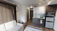 Located at 4106 12th Avenue NE in Seattle's vibrant University District, this thoughtfully designed studio offers a smart, modern layout that maximizes every inch of its single-floor footprint. The open-concept living and kitchen area anchors the space, featuring wood-look flooring throughout and a clean, contemporary aesthetic defined by crisp white walls and neutral tones.

The kitchen is well-appointed with two-tone cabinetry — dark lower cabinets paired with high-gloss white upper cabinets — a stainless steel sink, a compact refrigerator, a microwave, and track lighting above. The seamless countertop provides functional prep space, making the kitchen both practical and visually cohesive with the rest of the unit.

The living area accommodates a sofa, wall-mounted flat-screen TV, and a dedicated desk space, making it well-suited for both relaxation and work. A large window fills the room with natural light and offers views of the surrounding neighborhood.

A hallway connects the main living space to the bathroom and a neatly recessed in-unit stacked washer and dryer — a convenient and rarely found feature. The bathroom features light tile flooring, a dark wood vanity with a white countertop, and a glass-enclosed shower, all finished to a modern standard. The unit's efficient design, quality finishes, and practical amenities make it an excellent option in Seattle.