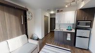 Located at 4106 12th Avenue NE in Seattle's vibrant University District, this thoughtfully designed studio offers a smart, modern layout that maximizes every inch of its single-floor footprint. The open-concept living and kitchen area anchors the space, featuring wood-look flooring throughout and a clean, contemporary aesthetic defined by crisp white walls and neutral tones.

The kitchen is well-appointed with two-tone cabinetry — dark lower cabinets paired with high-gloss white upper cabinets — a stainless steel sink, a compact refrigerator, a microwave, and track lighting above. The seamless countertop provides functional prep space, making the kitchen both practical and visually cohesive with the rest of the unit.

The living area accommodates a sofa, wall-mounted flat-screen TV, and a dedicated desk space, making it well-suited for both relaxation and work. A large window fills the room with natural light and offers views of the surrounding neighborhood.

A hallway connects the main living space to the bathroom and a neatly recessed in-unit stacked washer and dryer — a convenient and rarely found feature. The bathroom features light tile flooring, a dark wood vanity with a white countertop, and a glass-enclosed shower, all finished to a modern standard. The unit's efficient design, quality finishes, and practical amenities make it an excellent option in Seattle.