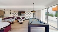 Located at 525 N Wilton Pl in Los Angeles, this single-floor community amenity space blends modern design with versatile social functionality. The open-plan layout seamlessly connects a fully equipped kitchen, a dining area, a lounge, and a billiards zone, creating an inviting environment for gatherings of all kinds.

The kitchen is a standout feature, outfitted with high-end stainless steel appliances, a full-size refrigerator, a gas cooktop, built-in ovens, and a large marble-topped island with bar seating. Wood-grain cabinetry and a striking marble backsplash contribute a refined, contemporary aesthetic. Pendant lighting adds a sophisticated finishing touch above the island.

Adjacent to the kitchen, a dining table with deep burgundy chairs provides ample seating for group meals or events. The lounge area features an eclectic mix of floral and velvet accent chairs arranged around a circular coffee table, anchored by a large wall-mounted television set against a bold, colorful geometric-patterned accent wall.

The billiards area anchors the space with a full-size pool table beneath dramatic black-and-gold pendant lights. Light hardwood flooring runs throughout, unifying the entire space. Expansive glass sliding doors flood the room with natural light and offer direct access to an outdoor area, visible in the background and ideal for extending social activities beyond the interior.