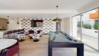 Located at 525 N Wilton Pl in Los Angeles, this single-floor community amenity space blends modern design with versatile social functionality. The open-plan layout seamlessly connects a fully equipped kitchen, a dining area, a lounge, and a billiards zone, creating an inviting environment for gatherings of all kinds.

The kitchen is a standout feature, outfitted with high-end stainless steel appliances, a full-size refrigerator, a gas cooktop, built-in ovens, and a large marble-topped island with bar seating. Wood-grain cabinetry and a striking marble backsplash contribute a refined, contemporary aesthetic. Pendant lighting adds a sophisticated finishing touch above the island.

Adjacent to the kitchen, a dining table with deep burgundy chairs provides ample seating for group meals or events. The lounge area features an eclectic mix of floral and velvet accent chairs arranged around a circular coffee table, anchored by a large wall-mounted television set against a bold, colorful geometric-patterned accent wall.

The billiards area anchors the space with a full-size pool table beneath dramatic black-and-gold pendant lights. Light hardwood flooring runs throughout, unifying the entire space. Expansive glass sliding doors flood the room with natural light and offer direct access to an outdoor area, visible in the background and ideal for extending social activities beyond the interior.