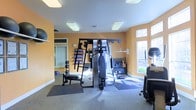 This two-floor space includes a dedicated fitness area designed to support a comprehensive workout experience. The gym room is finished with warm amber-toned walls that create an energizing atmosphere, complemented by neutral carpeted flooring and a clean white ceiling fitted with flush panel lighting that ensures the space is well-lit throughout. Multiple windows along the walls invite natural light and offer views of the surrounding greenery, contributing to an open and airy feel.

The fitness area is equipped with a multi-station cable and pulley strength training machine positioned centrally, serving as the focal point of the room. Along the walls, a vertical storage rack organizes a full set of dumbbells and weight plates, while a step and mat storage system keeps accessories neatly arranged. A weight bench with barbell equipment is also present, offering options for both free weight and machine-based training. Rubber floor mats are placed strategically beneath equipment to protect the flooring and provide stability during use.

The layout provides ample open floor space for stretching, functional movement, or additional equipment. The combination of natural and artificial lighting, thoughtful equipment arrangement, and vibrant wall color makes this a functional and inviting fitness environment suitable for a wide range of exercise routines.