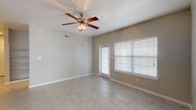 This single-floor space offers a clean, functional layout spread across several well-defined areas. The main living area is an open, airy room finished with neutral gray-toned tile flooring that runs consistently throughout the unit. A five-blade ceiling fan with a light fixture is centrally mounted, providing both air circulation and ambient lighting. A glass-paneled exterior door and a large window fitted with white horizontal blinds allow natural light to fill the space while offering access to the outdoors.

Additional rooms, likely bedrooms, mirror a similar aesthetic with the same tile flooring, ceiling fans, and windows dressed in matching white blinds that provide views of surrounding greenery. The rooms offer a neutral, move-in-ready canvas with light gray walls and white baseboards throughout.

The kitchen features warm-toned wood cabinetry paired with light granite-style countertops and a full suite of black appliances, including a range, over-the-range microwave, and refrigerator. A doorway from the kitchen leads to a dedicated laundry area. The bathroom complements the kitchen's design language, featuring a single-basin vanity with matching wood cabinetry, granite countertop, and a large mirror with overhead lighting.