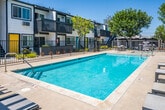 Located at 126 E 18th St in Costa Mesa, California, this single-story outdoor common area serves as the centerpiece of a contemporary residential community. The space is defined by a generously sized swimming pool with crystal-clear turquoise water, offering ample room for recreation and relaxation. Surrounding the pool, a spacious concrete deck is furnished with sleek lounge chairs, providing comfortable spots to enjoy the abundant Southern California sunshine.

Adjacent to the pool, a well-appointed spa offers a more intimate setting for unwinding, complemented by a nearby patio area with a dining table and chairs — ideal for outdoor gatherings. A clean, modern cabana structure with a pergola roof and a striped umbrella adds a stylish focal point to the poolside environment.

The community buildings framing the space feature a bold contemporary aesthetic, with white and dark-toned siding accented by vibrant, color-blocked doors in yellow, red, and teal. Upper-level units include private balconies that overlook the courtyard, while mature shade trees throughout the space contribute natural warmth and greenery. Black iron fencing delineates the pool area, reinforcing both safety and the cohesive modern design. The overall ambiance is bright, open, and inviting — well-suited to the Southern California lifestyle.