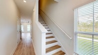 Located at 18 Sleeping Fox Run in Santa Rosa Beach, Florida, this two-story home offers three bedrooms, three bathrooms, and a thoughtfully designed layout spread across two floors. The main level features an open-concept kitchen equipped with white shaker cabinetry, granite countertops, a center island with sink, stainless steel appliances, and recessed lighting — all set atop light-toned hardwood-style flooring that flows throughout the home. The adjacent living area is bright and airy, benefiting from multiple windows and a ceiling fan, with a back door providing outdoor access. A hallway and attached garage with automatic opener round out the ground floor. A staircase with warm wood treads and crisp white risers connects the levels, leading to the upper floor where all three bedrooms are located. The spacious primary bedroom includes a dedicated closet with wire shelving systems, while the additional bedrooms offer natural light through windowed views of the surrounding greenery. The three bathrooms feature tile flooring, granite vanity tops, and tub-shower combinations with brushed bronze fixtures. Clean neutral walls and consistent flooring throughout lend the home a cohesive, move-in-ready aesthetic.