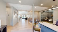 Located at 2500 McArthur Landing Cir in Fayetteville, NC, this single-floor community clubhouse offers a thoughtfully designed gathering space with a modern, mid-century aesthetic. Light hardwood flooring runs throughout, complemented by a neutral palette of warm beiges and soft blues that create a welcoming, cohesive ambiance.

The main living area serves as the heart of the space, anchored by a sleek linear fireplace topped with a wall-mounted television and a wooden mantel. Generous seating — including a deep teal sofa, rounded accent chairs, and leather armchairs — is arranged around a central coffee table, while large windows allow abundant natural light and views of an outdoor pool area. Adjacent to this zone, a billiards table and a stylish wet bar with counter seating provide additional entertainment options, set against a bold botanical-print accent wall.

A secondary lounge area features a slatted wood partition wall that adds architectural warmth and visual separation. Rounded upholstered chairs surround a low round table, and a glass-paneled sliding door leads into a private conference or co-working room equipped with a built-in desk and cabinetry. The entry corridor is accented with a curated gallery wall, a decorative console, and a gold-framed mirror, setting an inviting tone from the moment of arrival.