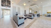 The Estero Oaks Luxury Living clubhouse is a single-floor community gathering space designed with a refined, contemporary aesthetic throughout. The main lounge area is expansive and light-filled, featuring soaring ceilings, recessed lighting, and large-format tile flooring laid in a classic diagonal pattern. Plush gray sofas and a sleek coffee table anchor the central seating area, creating an inviting atmosphere for residents to relax or socialize.

Adjacent to the main lounge is a dedicated social media lounge, where a series of flat-screen displays are mounted beneath the Estero Oaks Luxury Living branding wall. This room is furnished with crisp white sofas and dark accent tables, accented with gold and gray throw pillows for a warm, polished look.

A secondary lounge nook features cream upholstered chairs arranged on a circular patterned rug, complemented by curated artwork on the walls and abundant natural light from nearby windows.

The clubhouse kitchenette is well-appointed with dark cabinetry, stone countertops, pendant lighting, and a coffee station, making it ideal for community events. Floor-to-ceiling windows in the café-style dining area frame stunning views of the outdoor pool, pergola, and swaying palm trees, seamlessly connecting the interior to the tropical outdoor amenity space beyond.